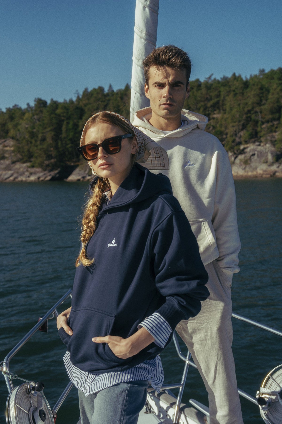 Two people on a sailing boat in the archipelago wearing The Gårdisto Hoodie in navy and sand.
