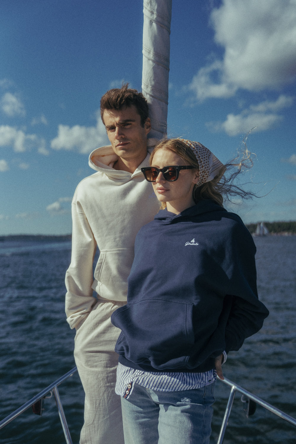 A couple on the bow of a sailing boat wearing The Gårdisto Hoodie in sand and navy.