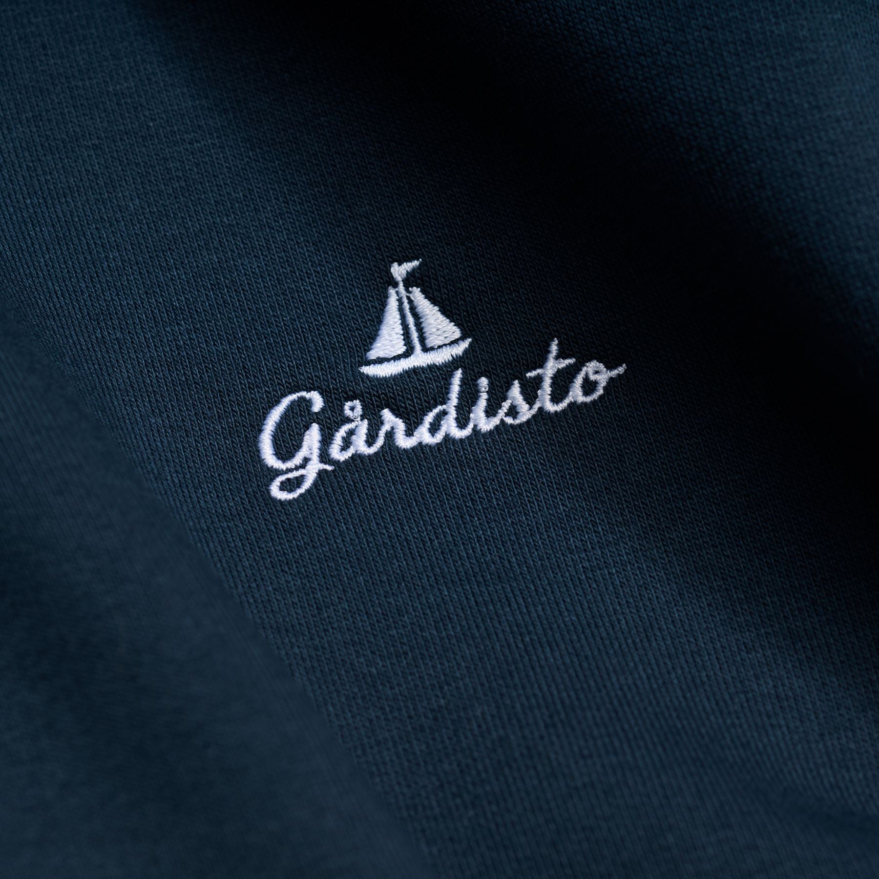 Nærbilde av Gårdisto-logoen med seilbåt og navnet brodert i hvit tråd på marineblått stoff